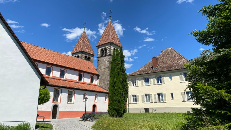 Basilika mit zwei Türmen umgeben von Bäumen und Grünflächen bei strahlend blauem Himmel.