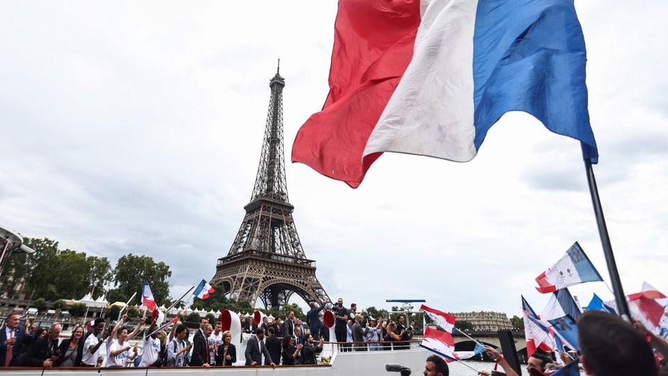 Fans und Athleten feiern und halten französische Nationalflaggen während einer Parade am Ufer der Seine.
