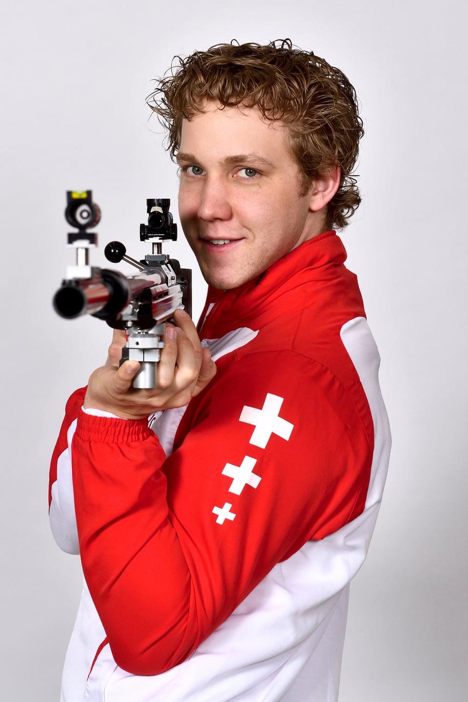 Junger Mann mit einem Sportgewehr in einem rot-weissen-Trainingsanzug mit Schweizer Kreuzen drauf