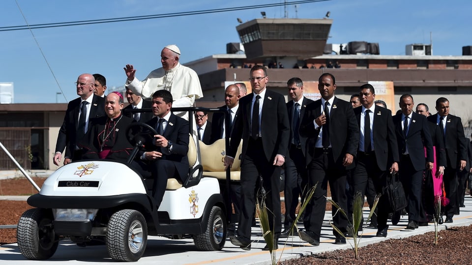 Papst stehend auf einem kleinen Auto, dahinter Männer in schwarzen Anzügen.