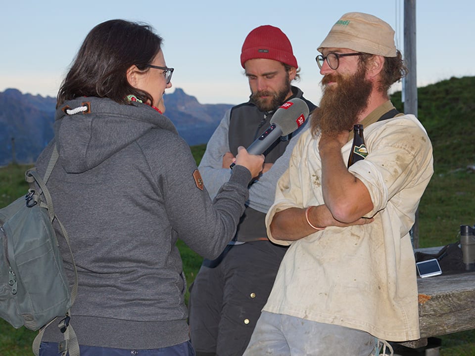 Eine Reporterin mit Mikrofon und zwei Männer auf einer Alp.