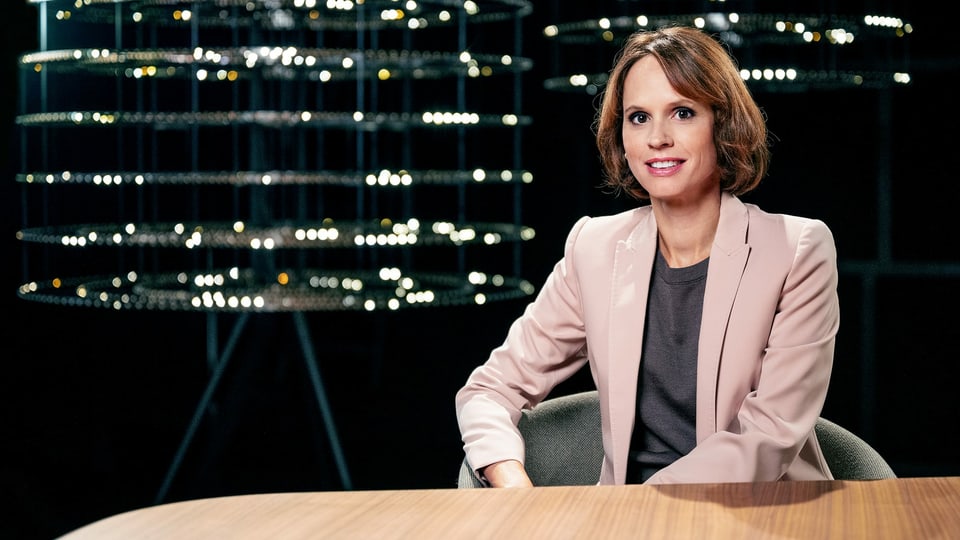 Frau in Blazer sitzt an einem Holztisch vor schwarzem Hintergrund