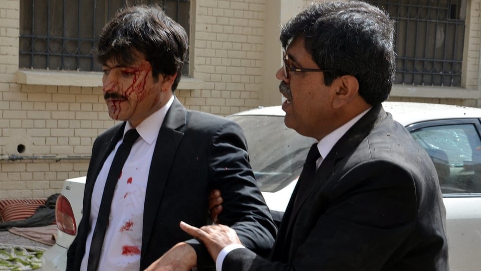Zwei Männer in Anzügen – einer blutet und wird von einem anderen am Arm geführt.