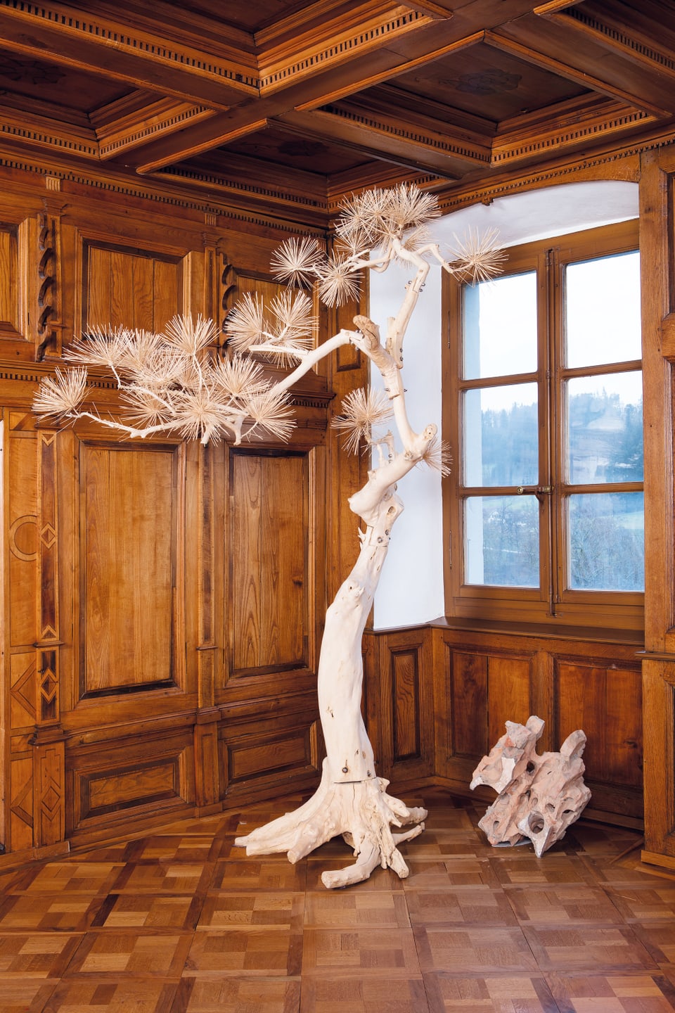 In einem getäferten Raum ist ein aus Holz hergestellter Pinienbaum zu sehen. Daneben ein weiteres, kleineres Objekt auf dem Boden. 
