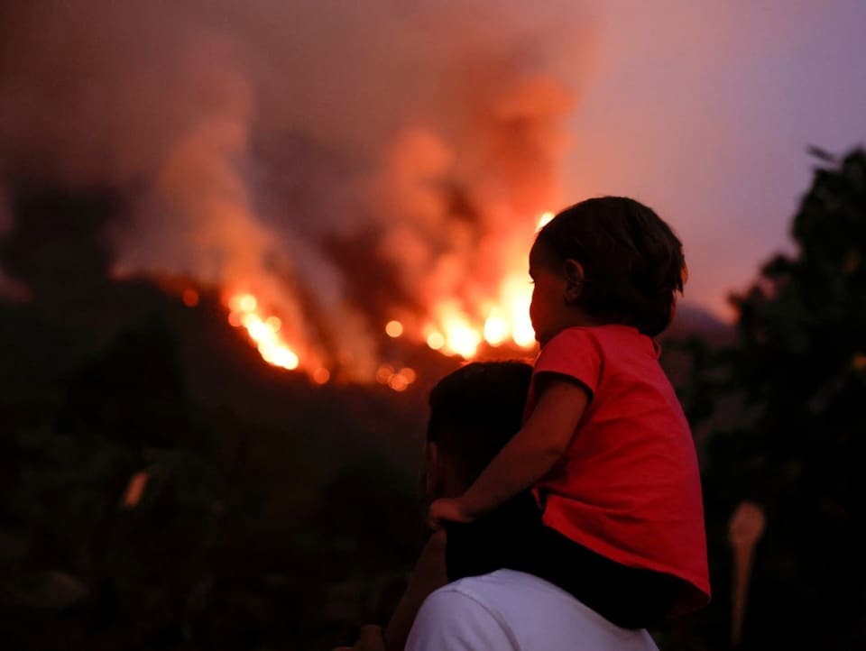 Ein Kind schaut auf den Schultern des Vaters auf den Waldbrand.