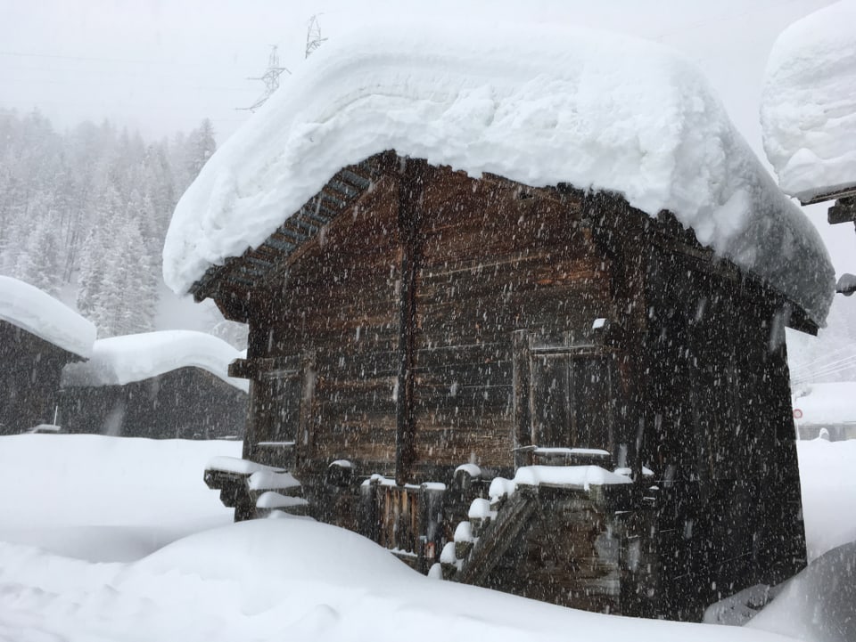 Holzhäuschen mit viel Neuschnee auf dem Dach in tief winterlicher Landschaft. 