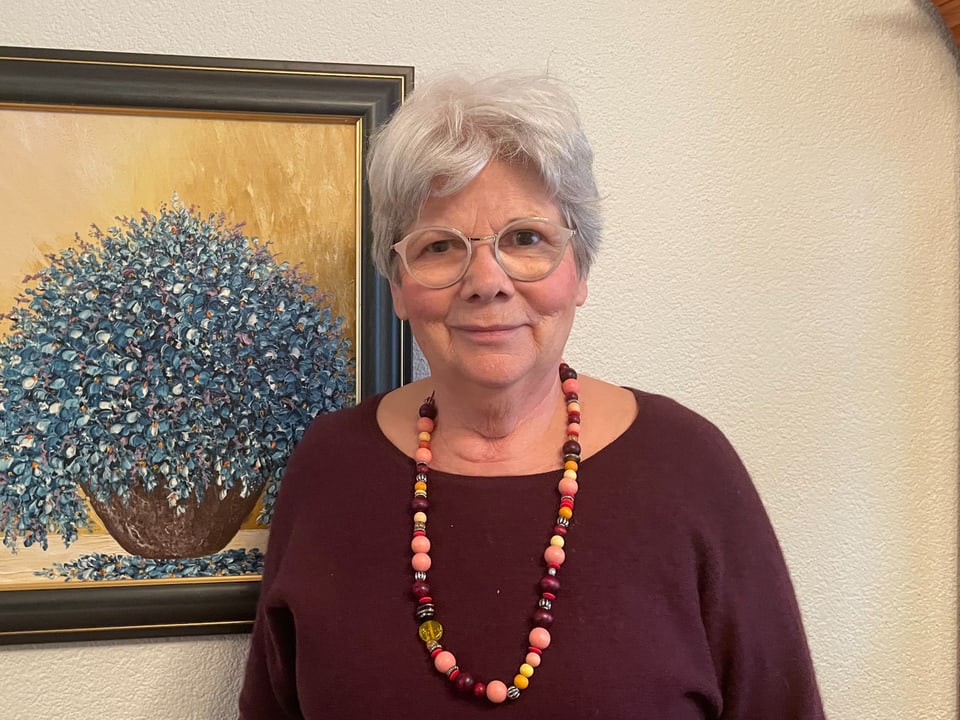 Ältere Dame mit grauen Haaren und bunten Halskette vor einem Gemälde.