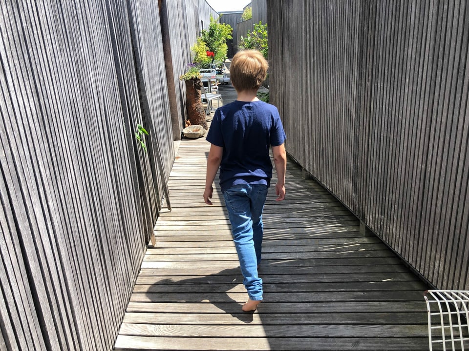 Ein Junge läuft  barfuss auf einem Holzsteg. Der Schatten ist sehr kurz.