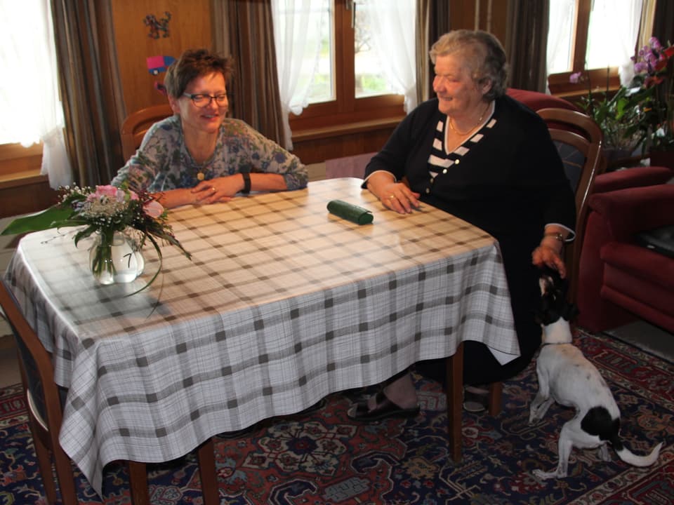 Zwei Frauen sitzen am Stubentisch. Eine streichelt einen kleinen Hund.