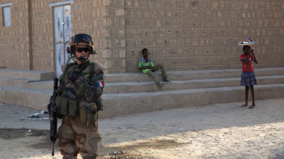 Ein Soldat in voller Kampfmontur mit gesenktem Maschinengewehr im Vordergrund. Dahinter zwei Kinder vor einer Hausmauer.