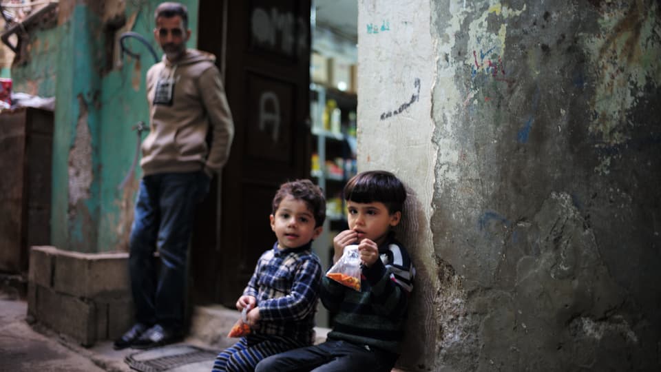 Zwei kleine Jungen sitzen vor einem Eingang in einem Flüchtlingslager. Einer schielt stark.