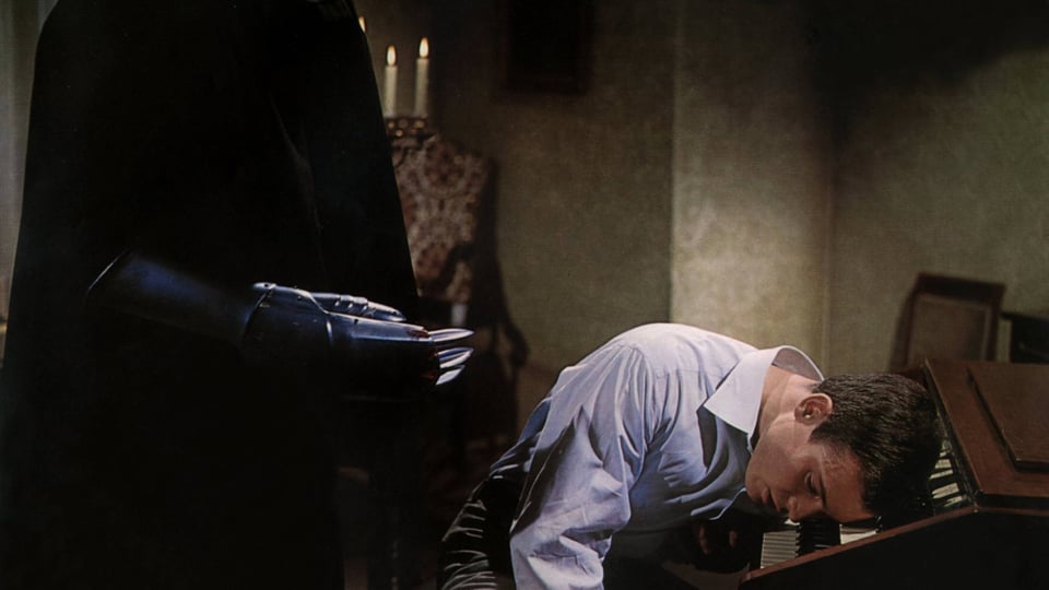 Ein Mann liegt mit seinem Kopf auf einem Klavier. Rechts von ihm eine Gestalt mit schwarzem Umhang und einer Eisenhand
