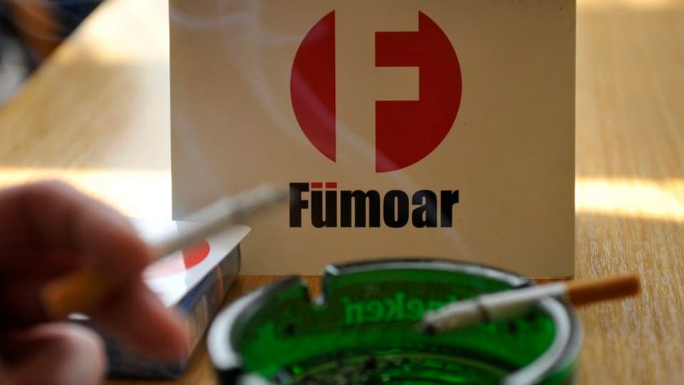 Ein grüner Aschenbecher mit einer brennenden Zigarette, dahinter ein weisses Schild mit der Aufschrift Fümoar.