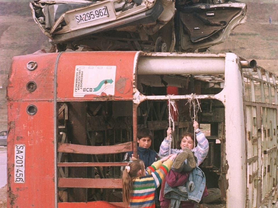 Drei Kinder spielen in einem Bus, der auf die Seite gekippt und zerstört ist.