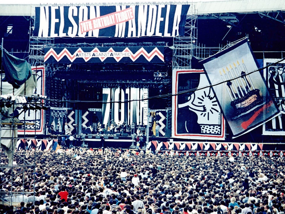 Tausende Zuschauer stehen vor einer grossen Bühne, über der in grossen Buchstaben der Name Mandelas prangt.