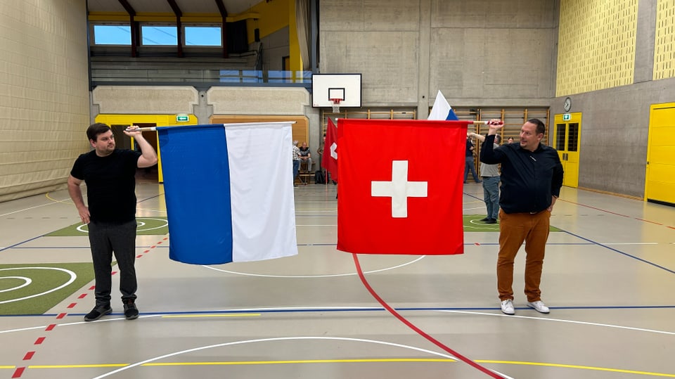 Zwei Männer stehen in Turnhalle und halten eine Schweizer- sowie eine Luzernerfahne waagerecht hoch.