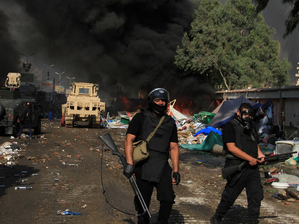 Zweil Polizisten in schwarzer Kleidung und mit Gasmaske. Im Hintergrund ein Militärfahrzeug und dichter, schwarzer Rauch.