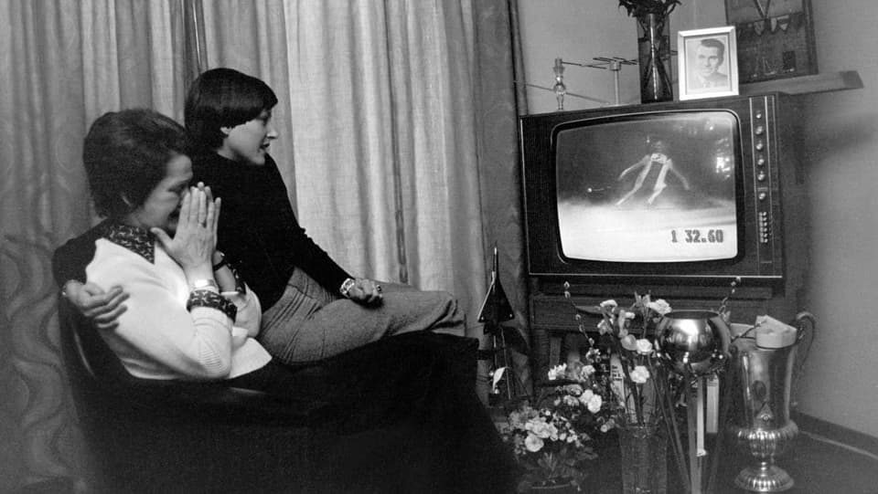 Schwarz-weiss-Aufnahme: Eine ältere und eine jüngere Frau sitzen gebannt vor dem Fernseher, in dem ein Skirennen läuft.
