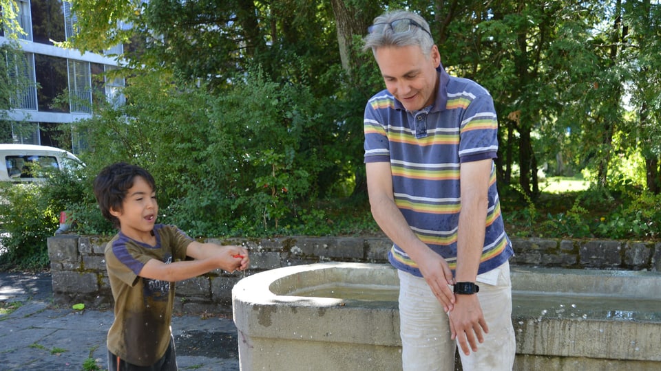Ein Mann und ein kleiner Junge vor einem Brunnen; der Junge spritzt Wasser.