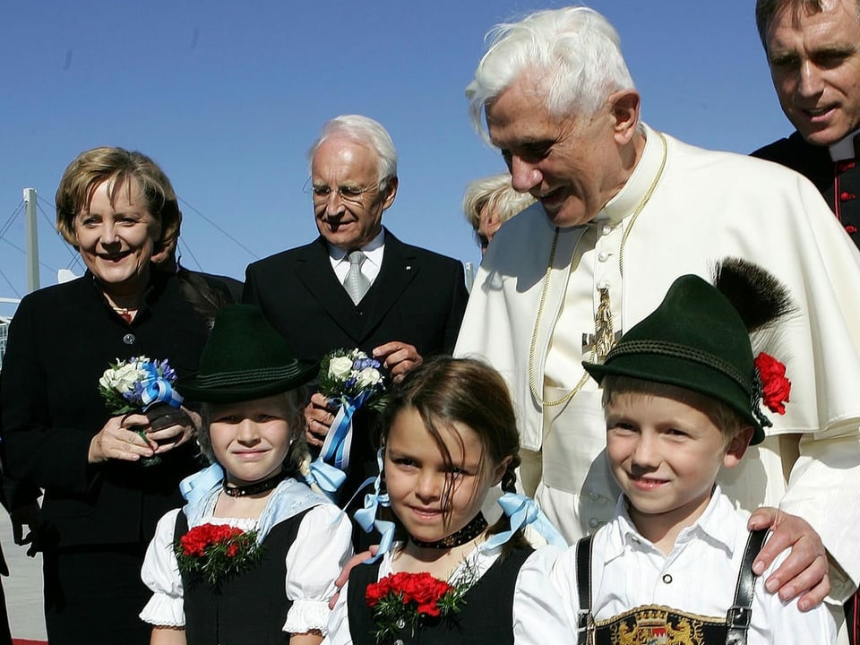 Papst Benedikt XVI. steht mit drei Kindern in traditioneller Kleidung neben Bundeskanzlerin Angela Merkel und dem bayerischen Landeshauptmann Edmund Stoiber.