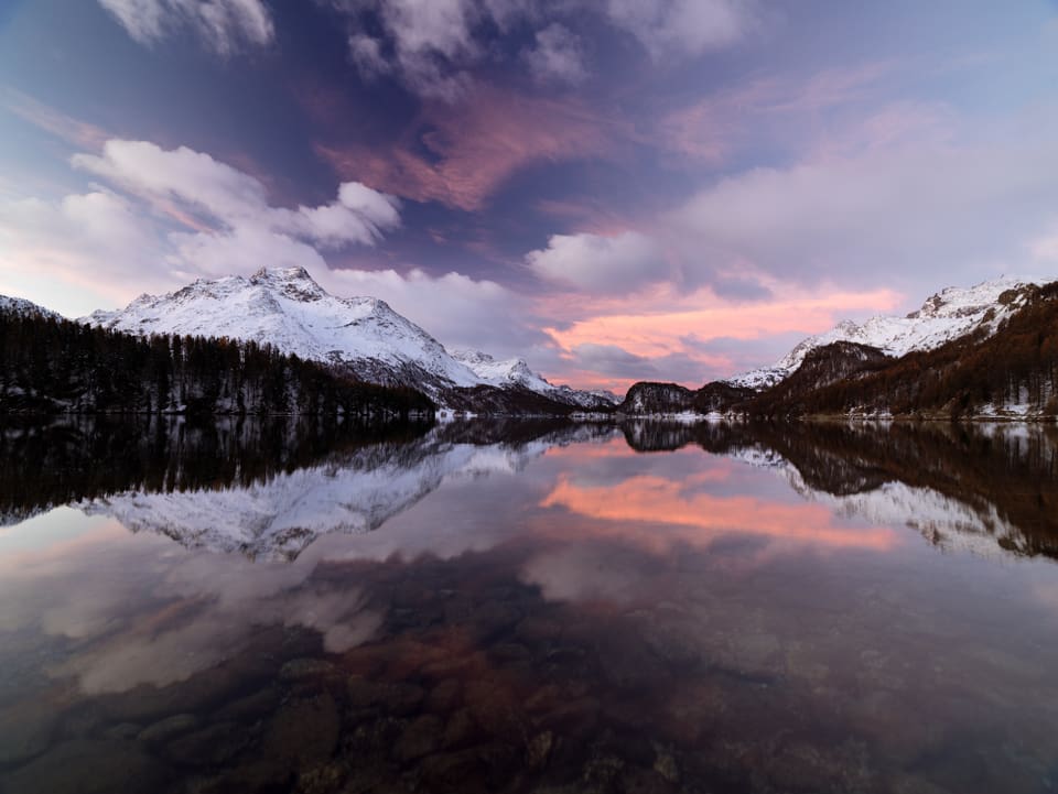 Im Vordergrund ein spiegelglatter Bergsee, in dem sich die verschneiten Talflanken und der Morgenhimmel spieglen. 