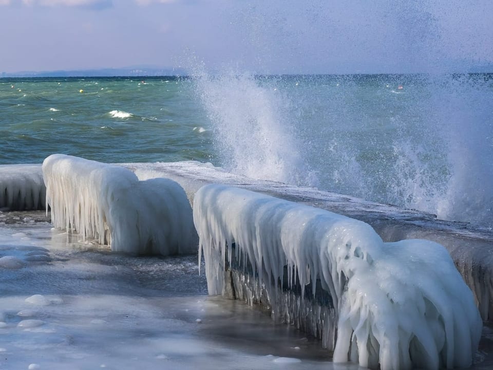Blick auf Seeufer mit eingefrorenen Sitzbänken. Dahinter sieht man Wellen und den Bodensee.