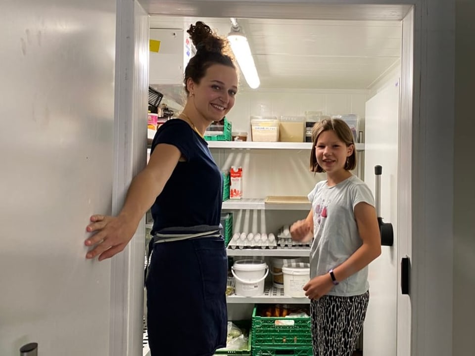 Links eine Frau in Uniform, rechts ein lächelndes Mädchen. Die beiden stehen in einem begehbahren Kühlschrank.