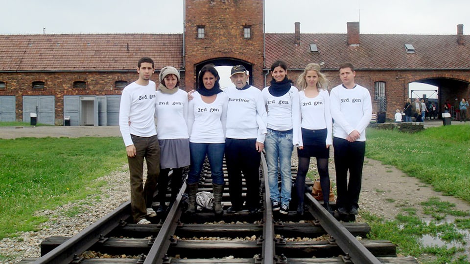 Sieben Menschen stehen auf den Geleisen in Auschwitz. Sie tragen T-Shirts, auf denen entweder "2nd gen" oder 3rd Gen" steht. Der Mann in der Mitte trägt ein T-Shirt, auf dem "Survivor" steht. 