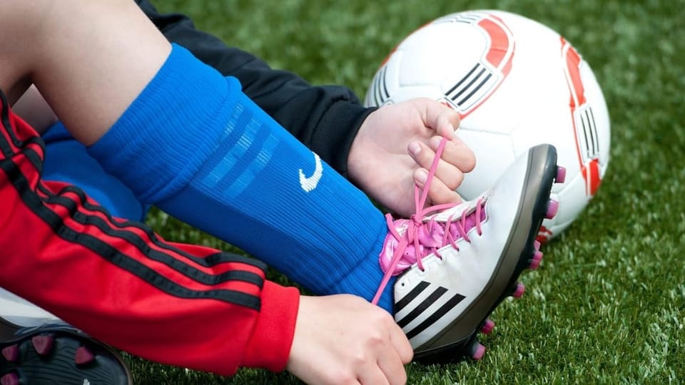 Kinderhände binden sich die rosa Schnürsenkel von Fussballschuhen. Daneben liegt ein Fussball.
