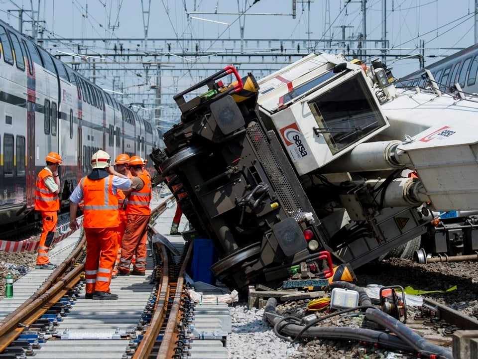 Arbeiter neben dem umgestürzten Kran und ein Zug, der an ihnen vorbeifährt.