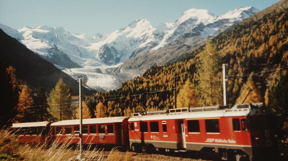 Ein roter Zug fährt durch eine Engadiner Landschaft, im Hintergrund der Morteratschgletscher.