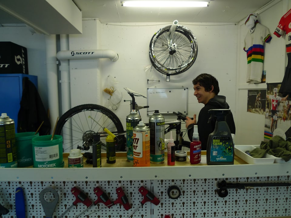 Zu sehen ist eine Velowerkstatt mit allerlei Werkzeugen an der Wand aufgehängt, Dosen, Gütterli und Flaschen auf einem Tisch und Velo-Trikots an der Wand.