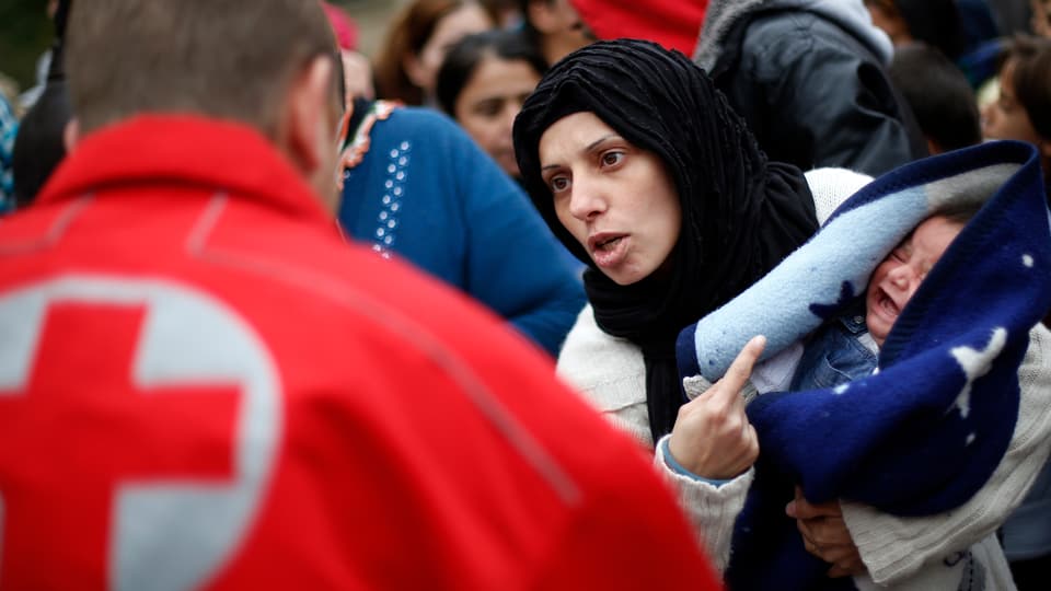 Eine syrische Frau zeigt auf ihr Baby auf dem Arm, im Vorderdrund ein Mann in einer Rot-Kreuz-Jacke.