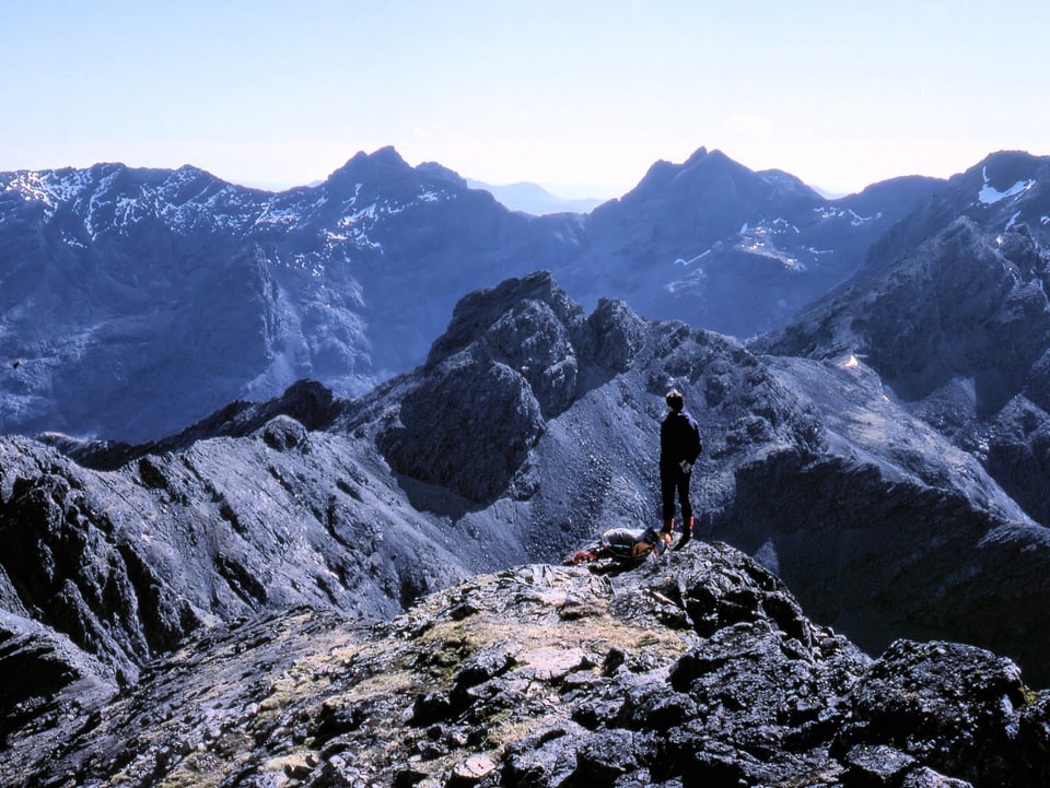 Eine Person auf einem Gipfel schaut auf schwarze, felsige Bergketten.