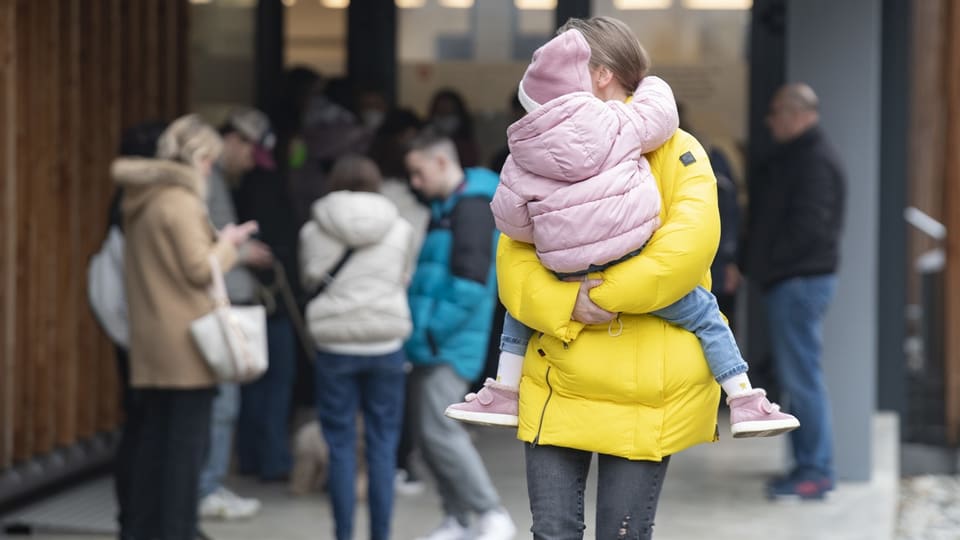 Frau mit gelber Jacke hält Kind mit rosaroter Jacke in den Armen.