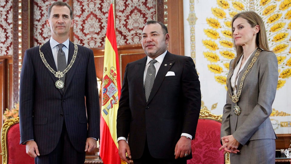 Felipe, Mohammed und Letizia stehen stramm vor roten Sesseln und einer spanischen Flagge.