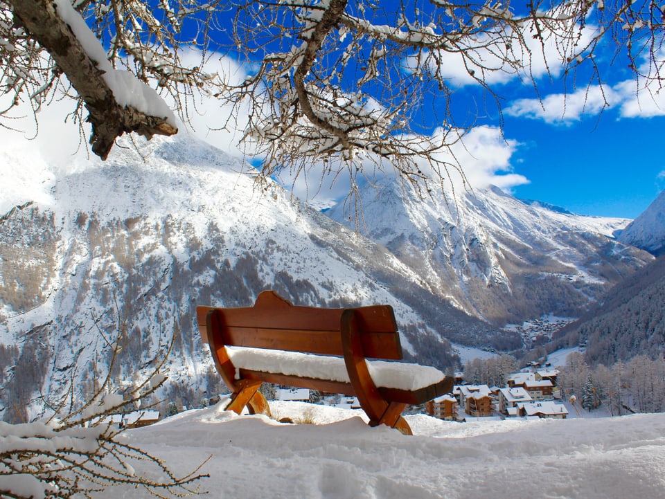 Eine frisch verschneite Sitzbank. Im Hintergrund Bergpanorama mit blauem Himmel.