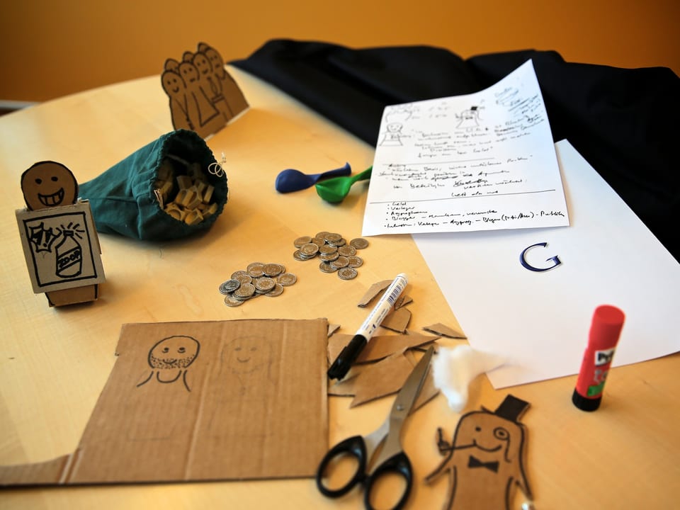 Pappmännchen, Kleingeld, Scrabble-Steine und Bastelmaterial liegen auf einem Bürotisch.