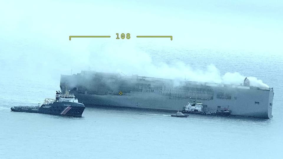 Ein graues Frachtschiff mit einer grossen Rauchwolke, daneben ein Schiff der niederländischen Küstenwache