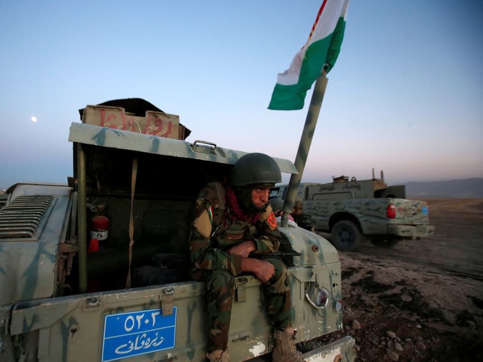Ein Mitglied der Peschmerga-Armee sitzt auf einem Militärfahrzueg, danaben die irakische Flagge.