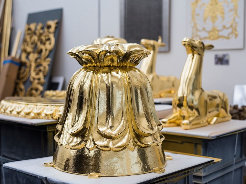 Glänzende, goldene Objekte in einem Atelier: Eine verzierte Glocke und eine Tierstatue.
