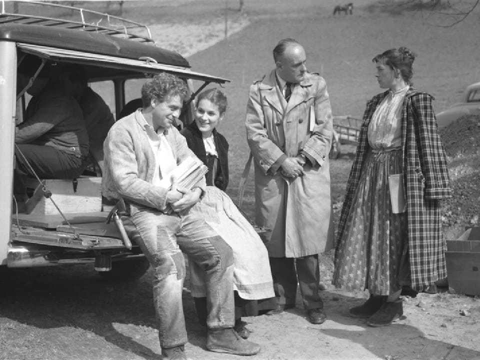 Uli und Vreneli sitzen auf der Heckklappe eines Auto. Neben ihnen stehen ein Mann und eine Frau.,
