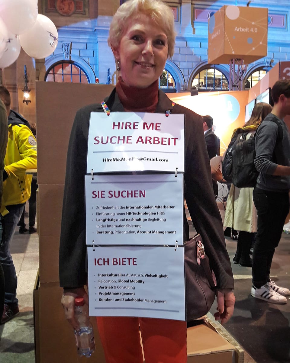 Eine ältere Dame steht am HB Zürich. Sie hat sich mit Schildern behängt, auf denen steht, dass sie eine Stelle sucht.