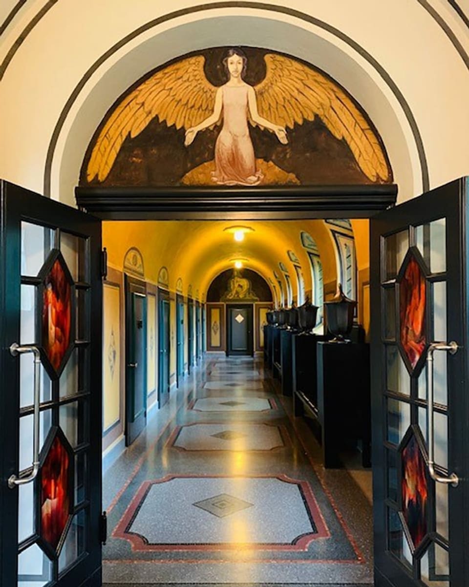 Der Eingang einer Aufbahrungshalle, warmes Licht, Mosaikböden, eine geöffnete Glasdoppeltüre. Über der Tür hat eine Wandmalerei, einen Engel mit goldenen Flügeln.