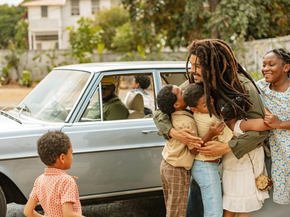 Ein Mann mit Rastas umarmt drei Kinder, eines steht vor ihnen. Im Hintergrund ein Auto mit Chauffeur.