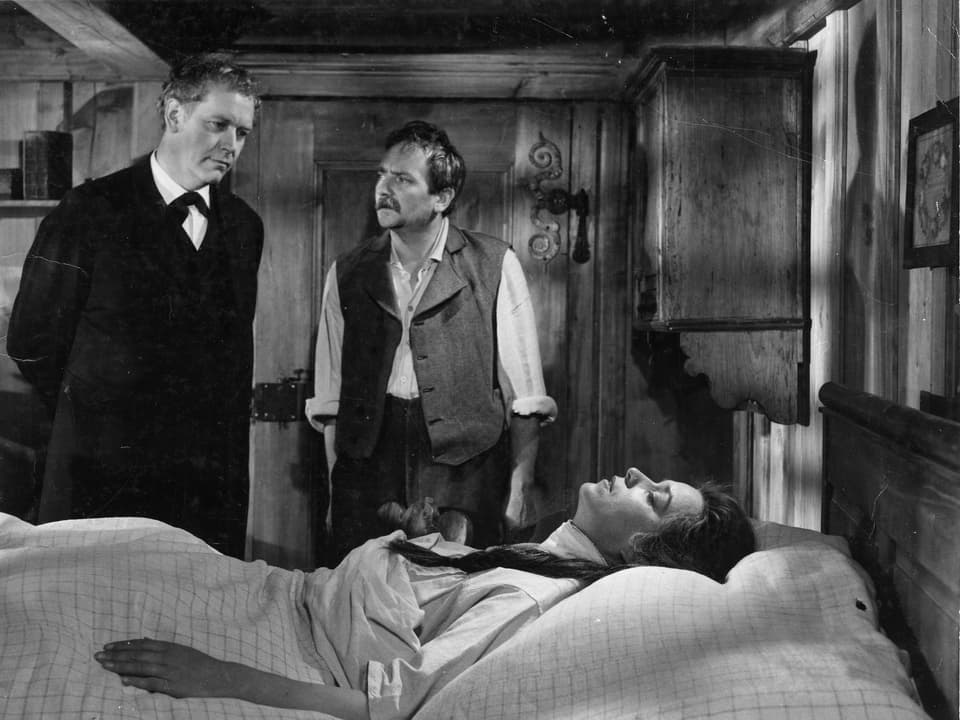 Eine Frau liegt mit geschlossenen Augen in einem Bett. Hinter dem Bett stehen zwei Männer. Einer von ihnen ist ein Arzt. Der andere schaut den Arzt fragend an.