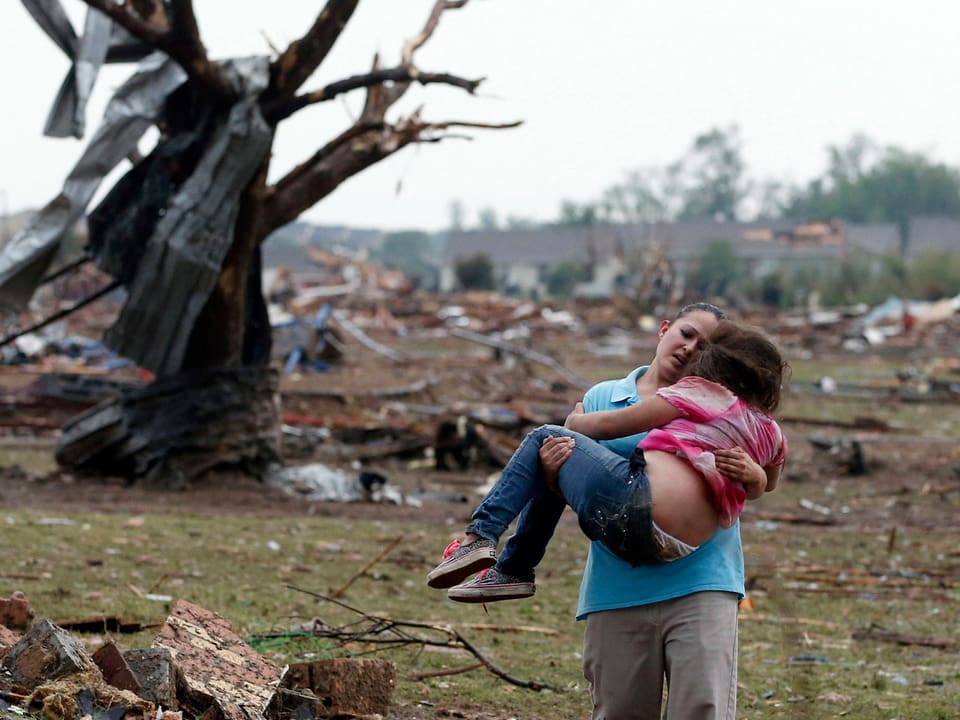 Eine Frau trägt ein Kind auf den Armen. Im Hintergrund Trümmer.