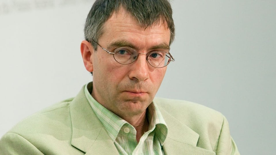 Ein Mann mit hellgrüner Jacke, kariertem Hemd, Brille und kurzen Haaren. 