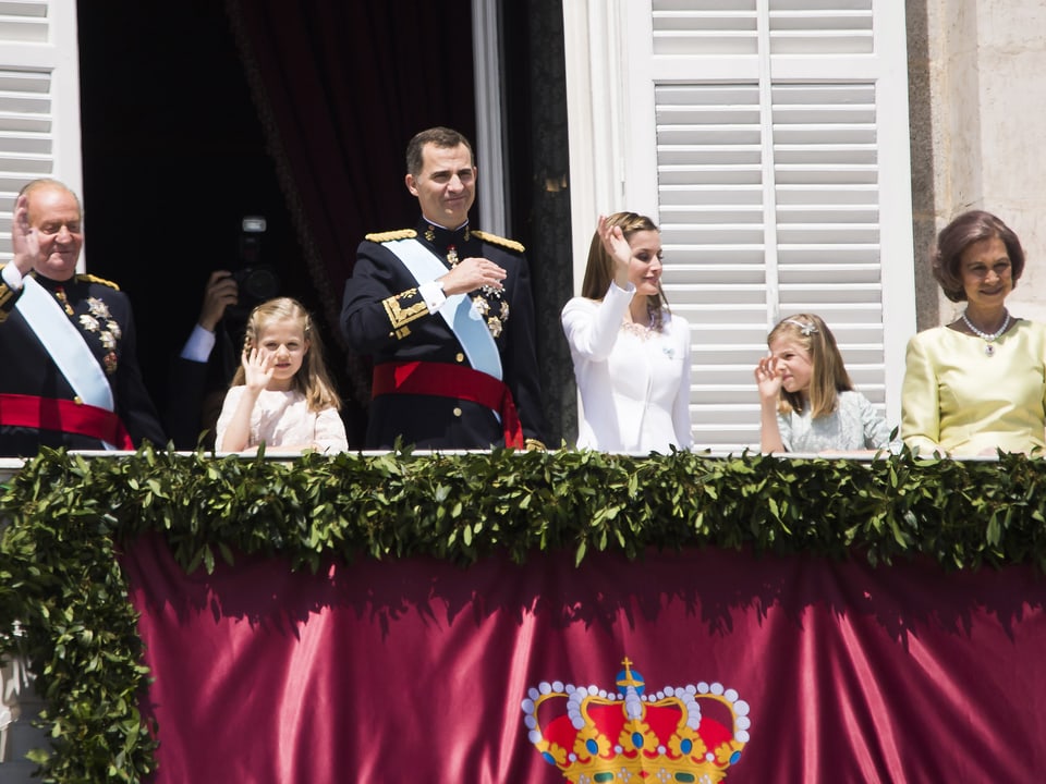 Royale Familie bestehend aus Vater, Grossvater, Enkelin, Frau und Grossmutter stehen auf einen geschmückten Balkon und winken.