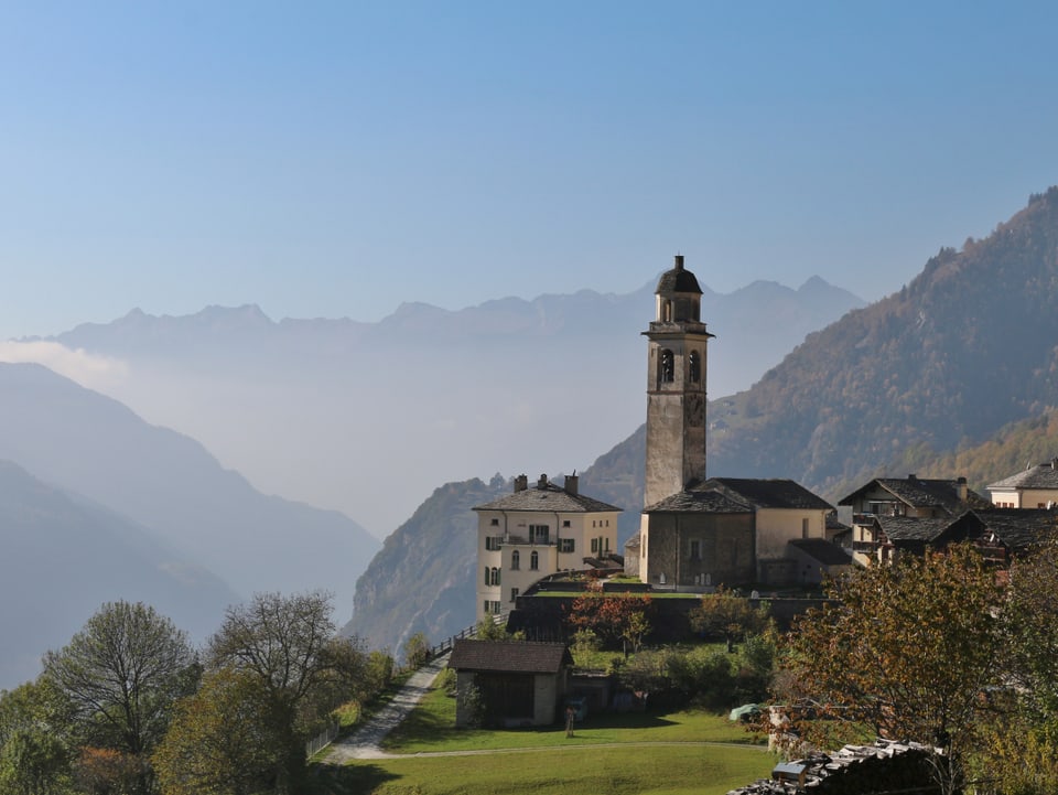 Im Vordergrund das Dorf Soglio mit der Kirche. Im Hintergrund ist es über dem Tal dunstig, oben ist der Himmel blau.